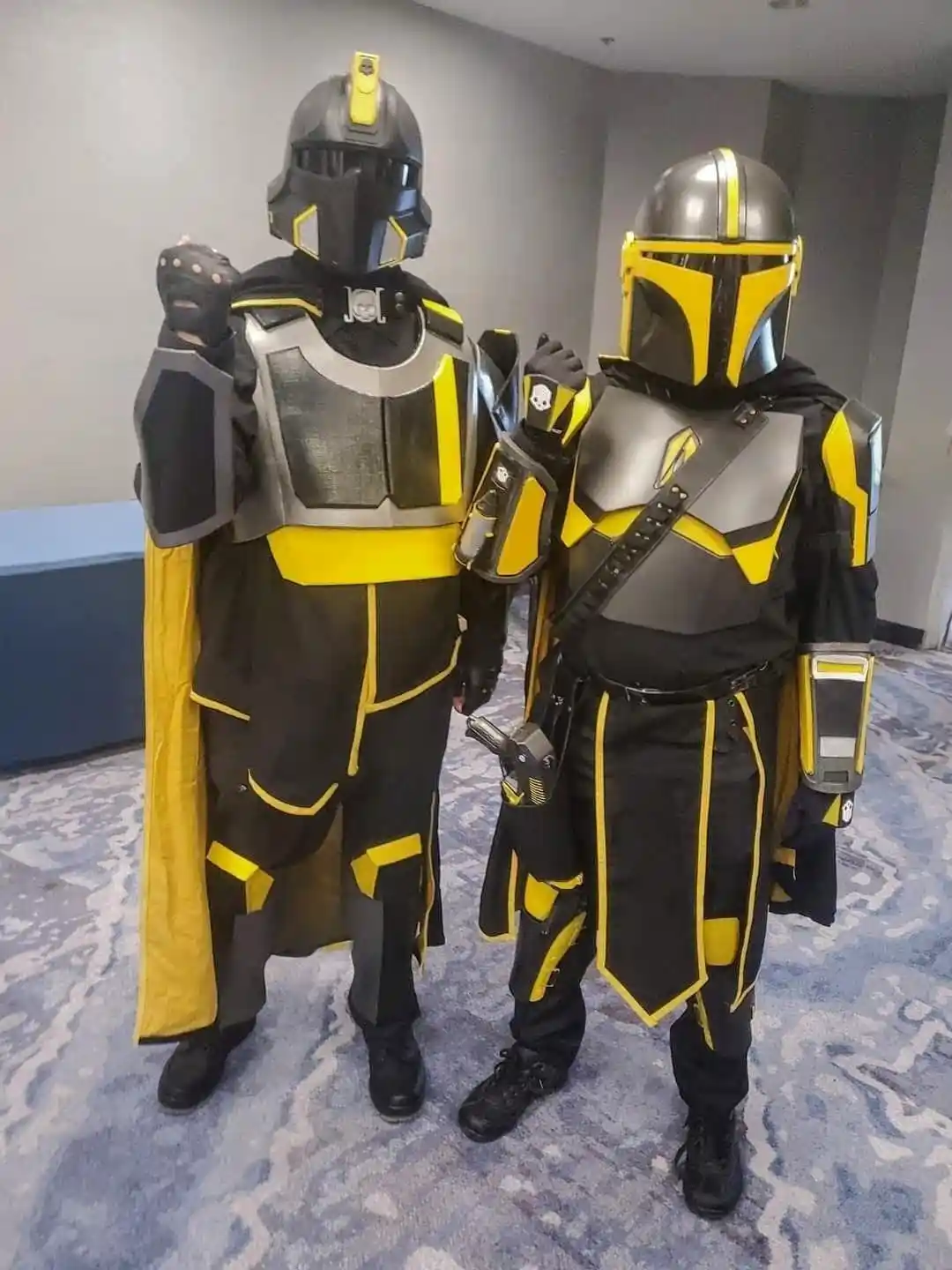 Two cosplayers stand side by side in a hotel hallway, both wearing detailed Mandalorian armor with black and yellow color…