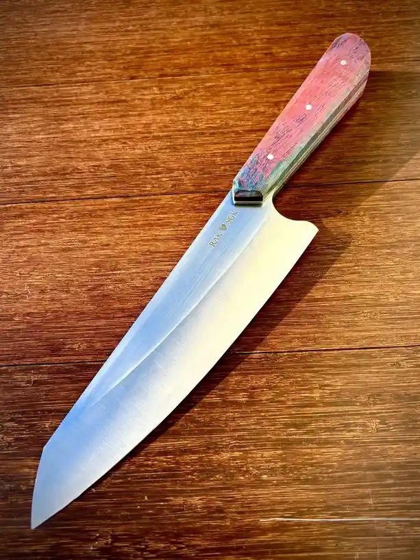 A close-up photo shows a chef's knife with a colorful, marbled pink and blue handle resting on a dark wooden surface. The…