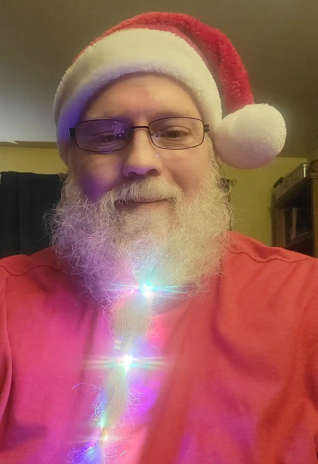 A man with glasses and a white beard wears a red and white Santa hat and a red shirt, smiling at the camera. A string of…