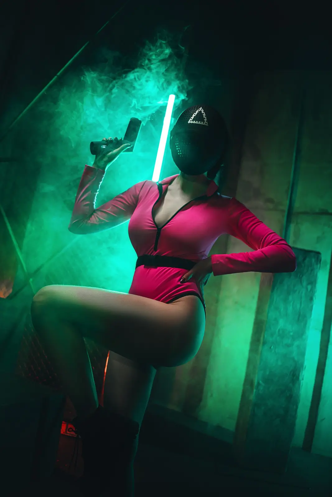 A person in a pink bodysuit and black mask with a white triangle emblem holds a glowing green lightsaber-like object while…
