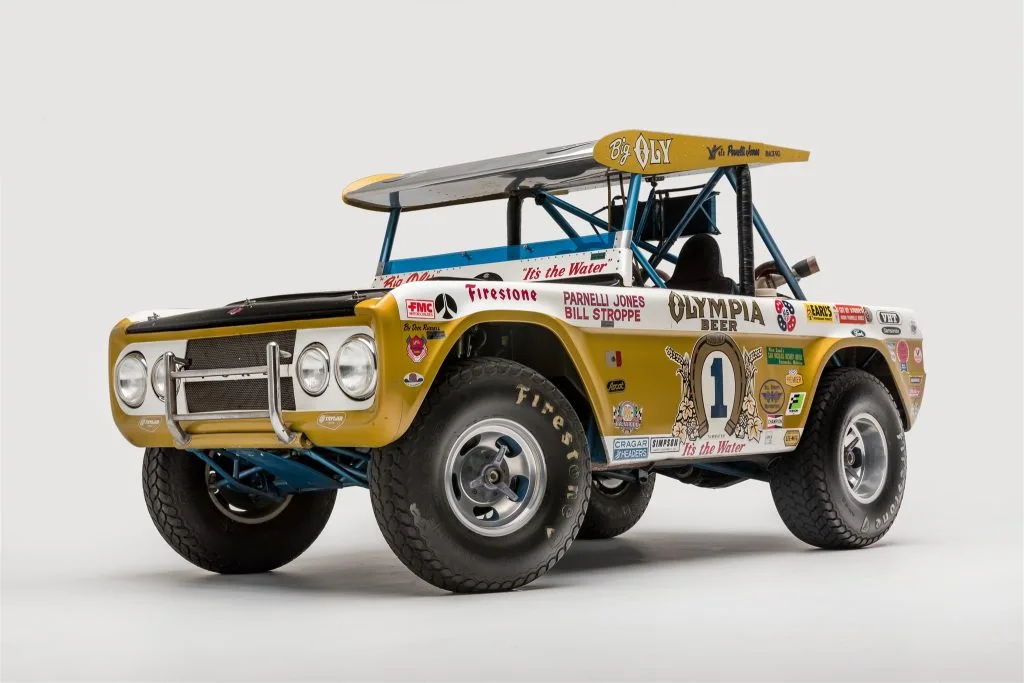 A yellow and white vintage off-road race truck with a roll cage, large tires, and numerous sponsor decals including…
