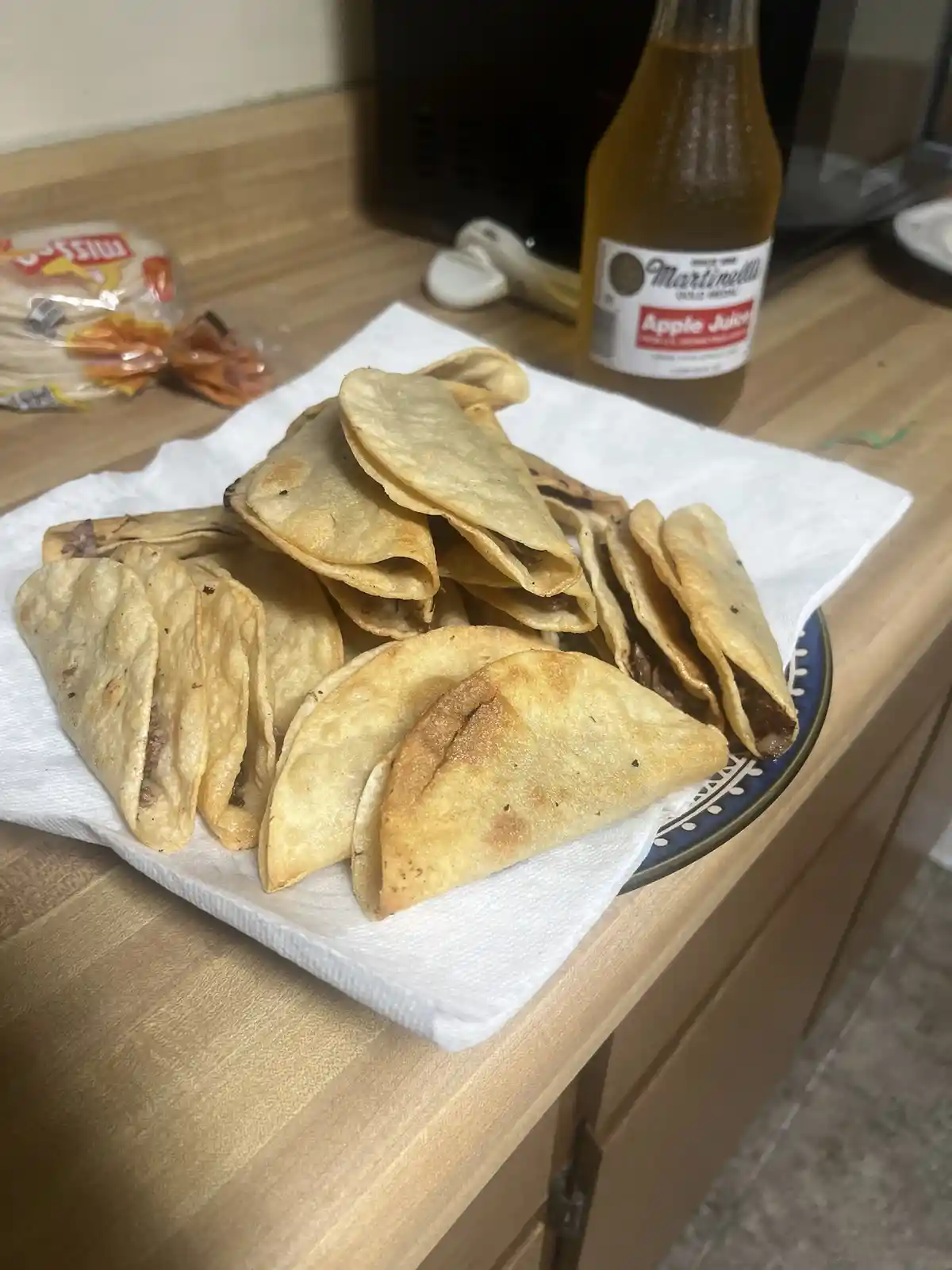 A plate of fried tacos sits on a paper towel on a wooden countertop, next to a bottle of Martinelli's Apple Juice and a…