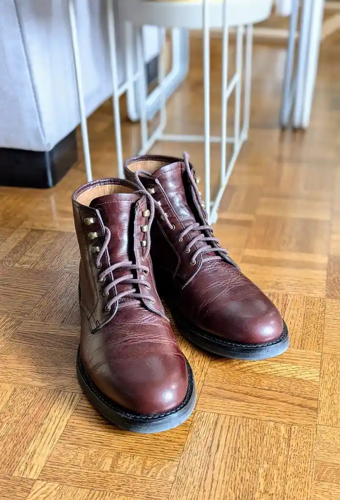 A pair of well-worn, burgundy leather ankle boots with metal eyelets and laces sit on a wooden parquet floor, with white…