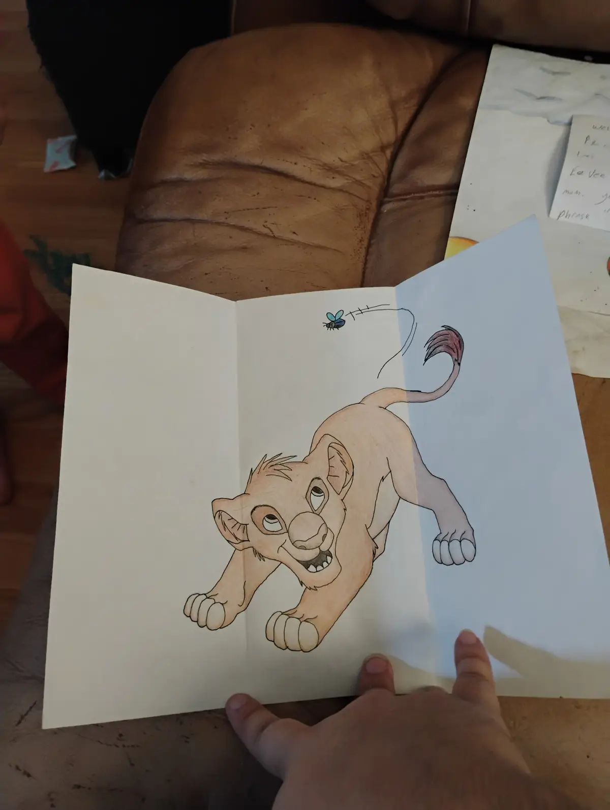 A hand holds open a piece of paper showing a colored drawing of a young lion cub, likely Simba from The Lion King, lunging…