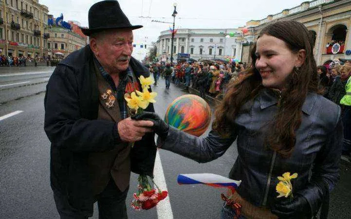 An elderly man with a black hat and military medals on his jacket receives yellow flowers from a smiling young woman holding…