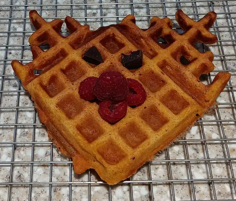 A golden-brown waffle rests on a metal cooling rack, with two chocolate pieces forming eyes and a cluster of raspberries…