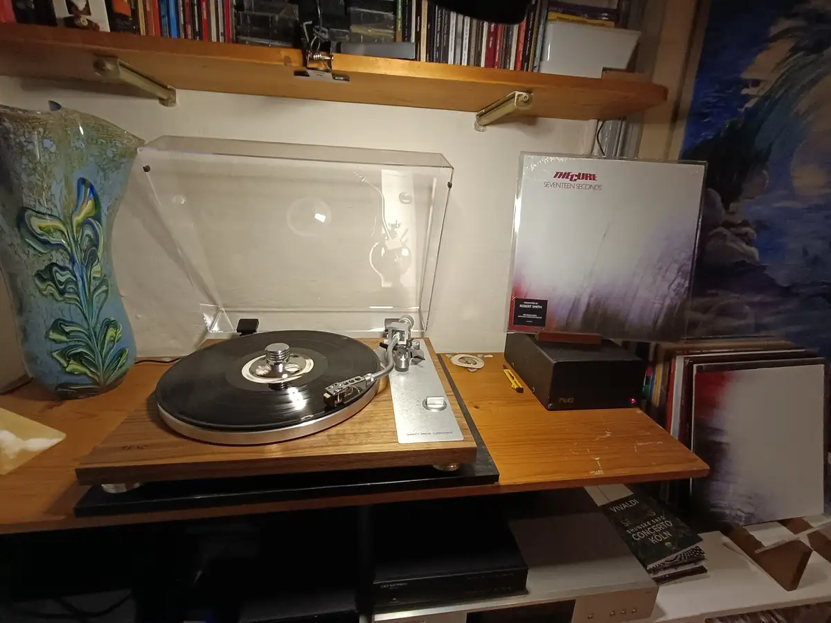 A vinyl record player sits on a wooden shelf with a clear dust cover, next to a sealed copy of The Cure's 'Seventeen…
