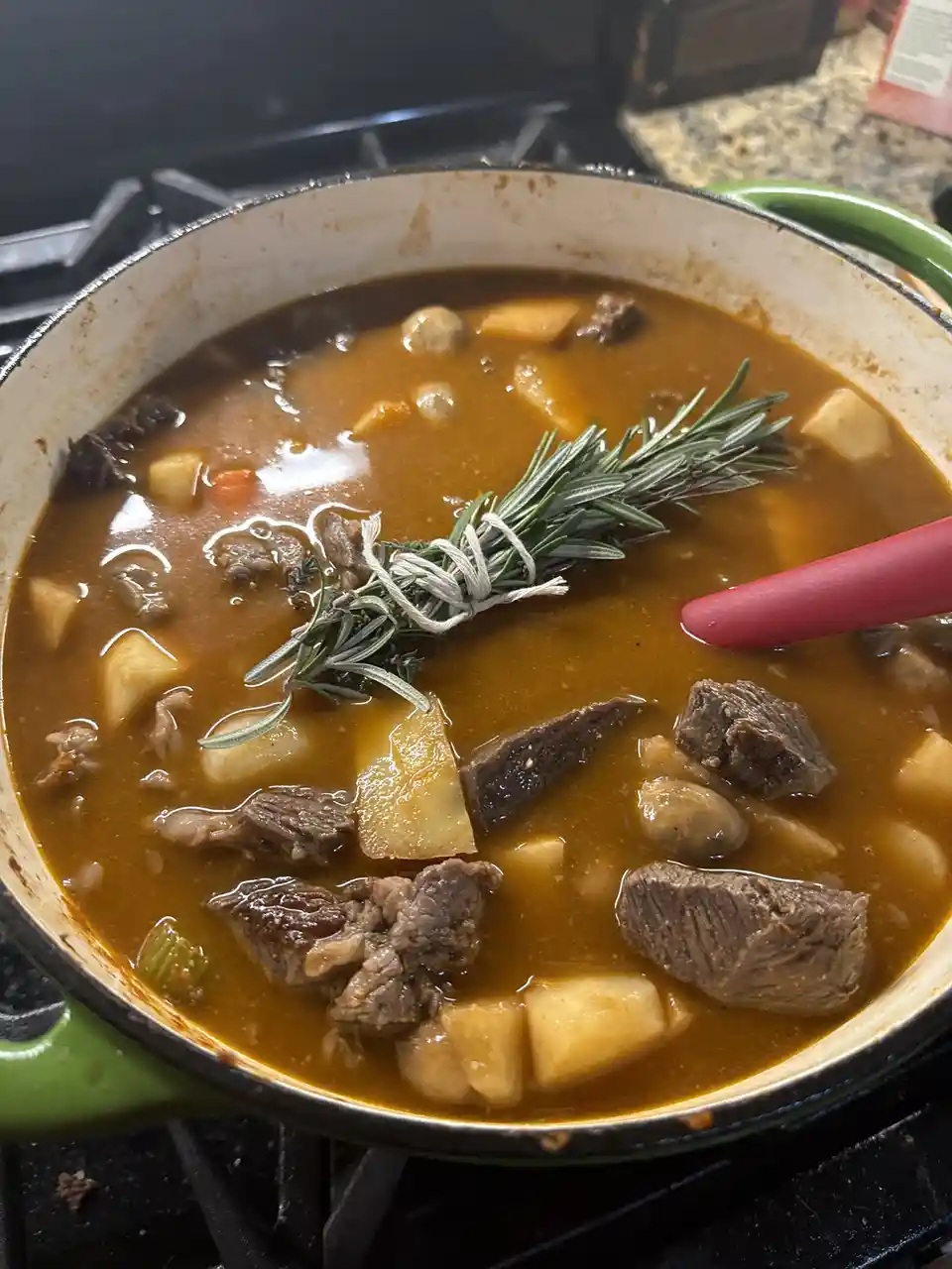 A close-up photo shows a green enameled cast iron pot on a stovetop, filled with a rich brown beef stew containing chunks of…