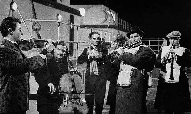 A black-and-white photo of five men in vintage attire playing string and brass instruments on a ship's deck at night. The…