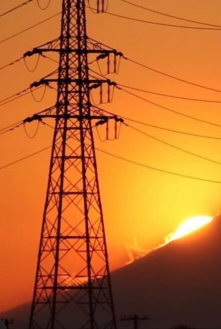 A silhouette of a tall electricity pylon stands against a vibrant orange and yellow sunset, with the sun partially obscured…