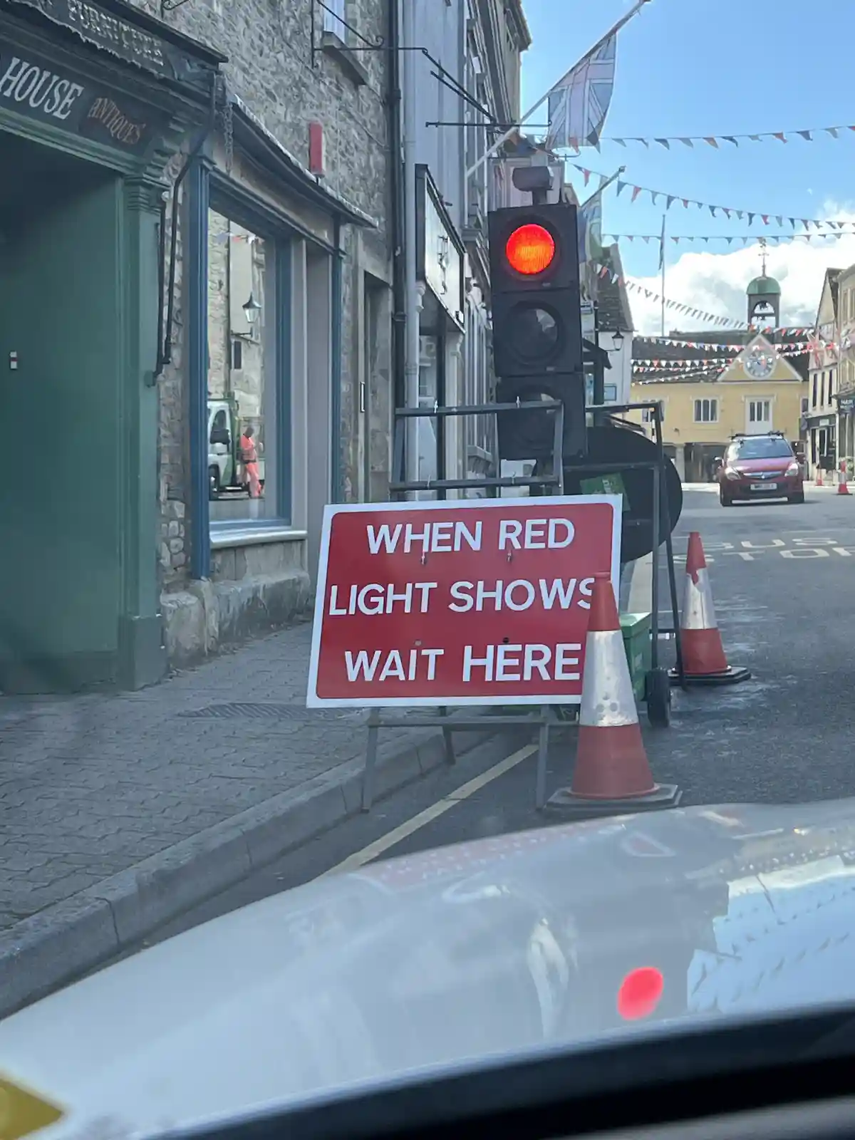 A street scene with a red traffic light and a red sign that reads 'WHEN RED LIGHT SHOWS WAIT HERE'. The photo is taken from…