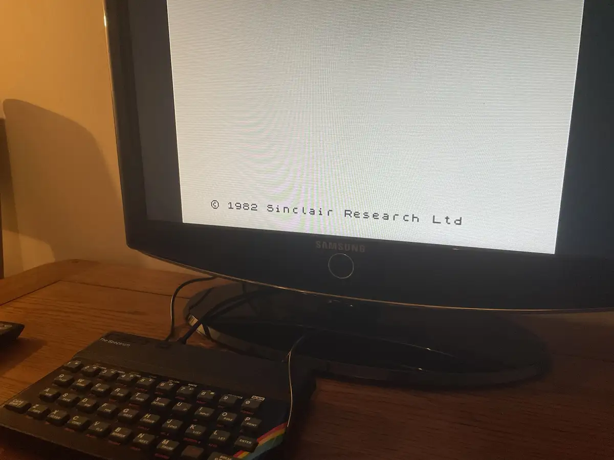 A modern Samsung monitor displays the vintage Sinclair ZX Spectrum startup screen with its copyright text, while a physical…