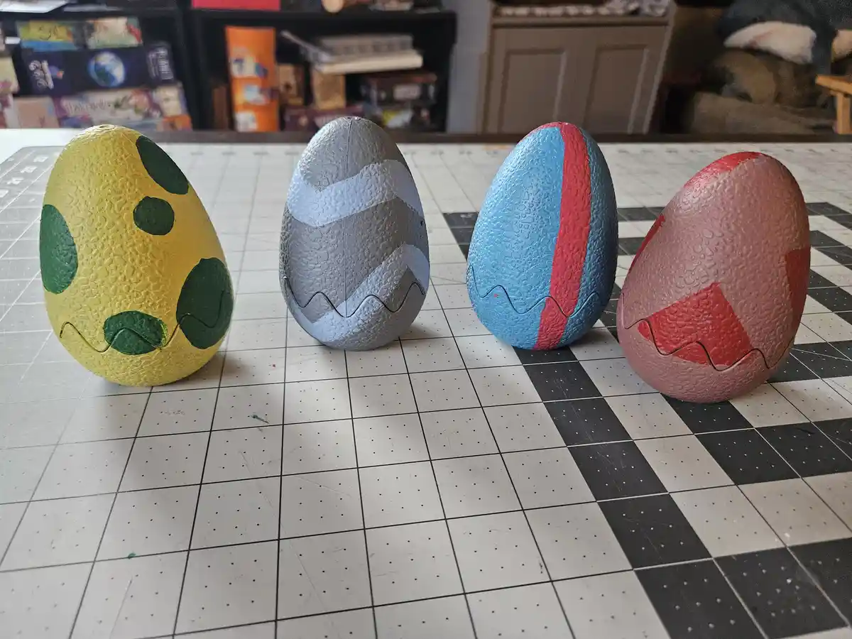 Four hand-painted, textured plastic eggs sit in a row on a checkered cutting mat. Each egg has a distinct color scheme and…