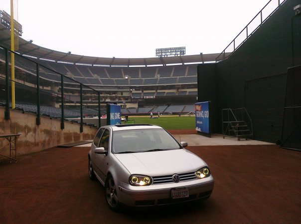 A silver Volkswagen Golf is parked on the dirt infield of a large, empty baseball stadium with bleachers and a green field…