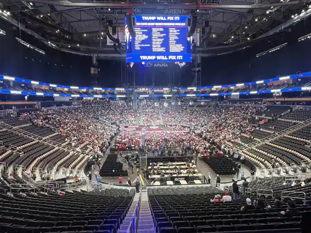 A wide-angle photo of a large, mostly empty arena with a crowd gathered near the stage. A massive central screen displays…