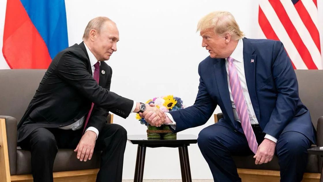 two men in suits, one with a russian flag behind him and one with an american flag, are shaking hands over a small table…
