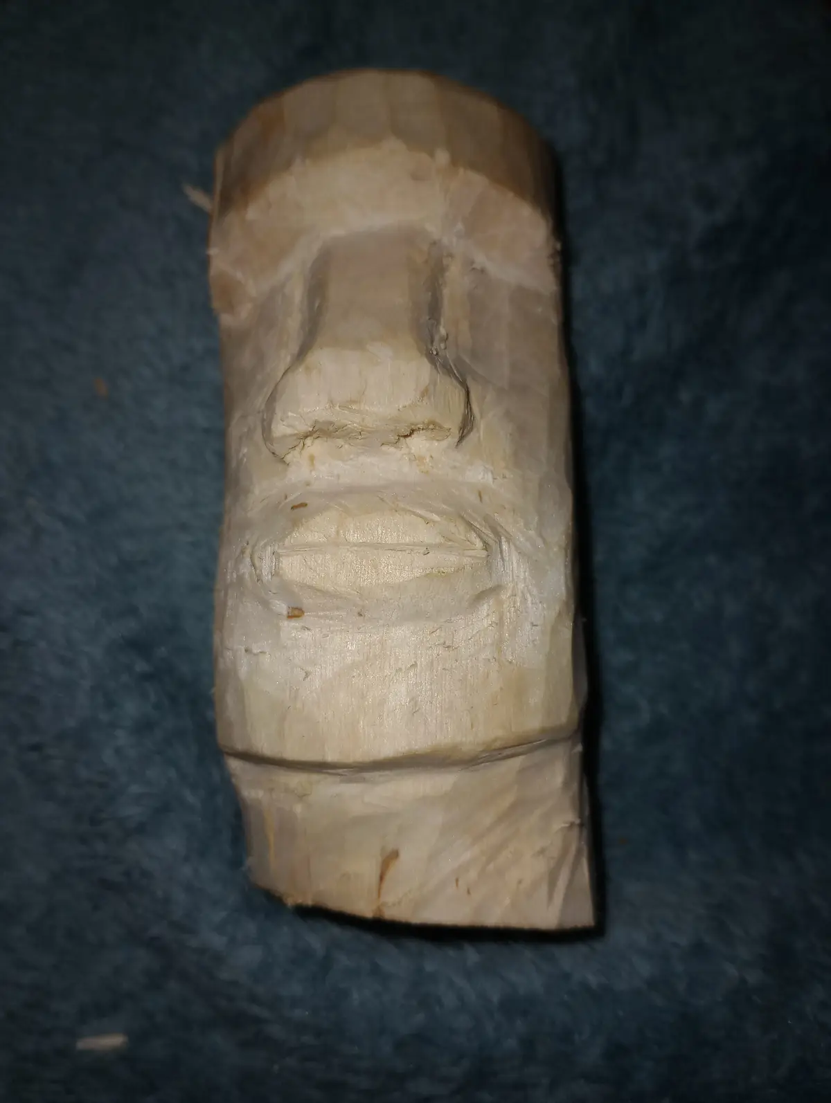 A rough, unfinished wooden carving of a Moai head sits on a dark blue textured surface, showing visible wood grain and…