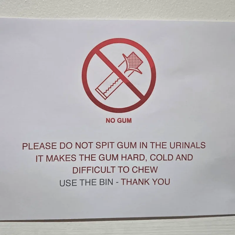 A white sign with red text and icon warns against spitting gum in urinals, explaining it makes gum hard, cold, and difficult…