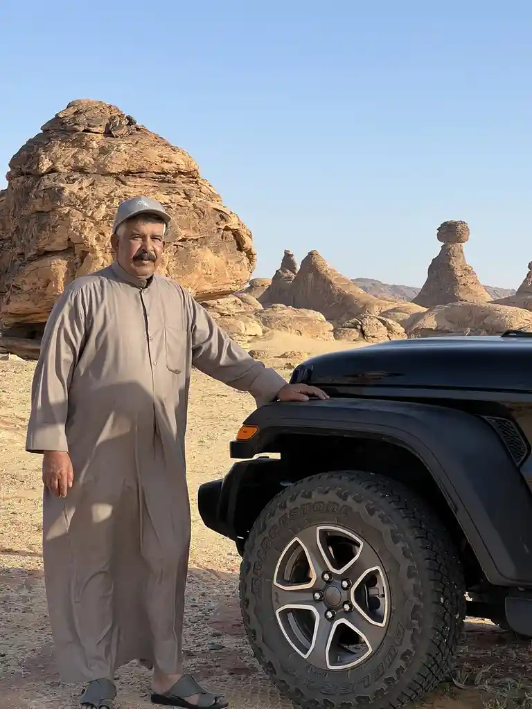 A man in traditional attire stands confidently next to a black Jeep in a desert landscape with distinctive rock formations…