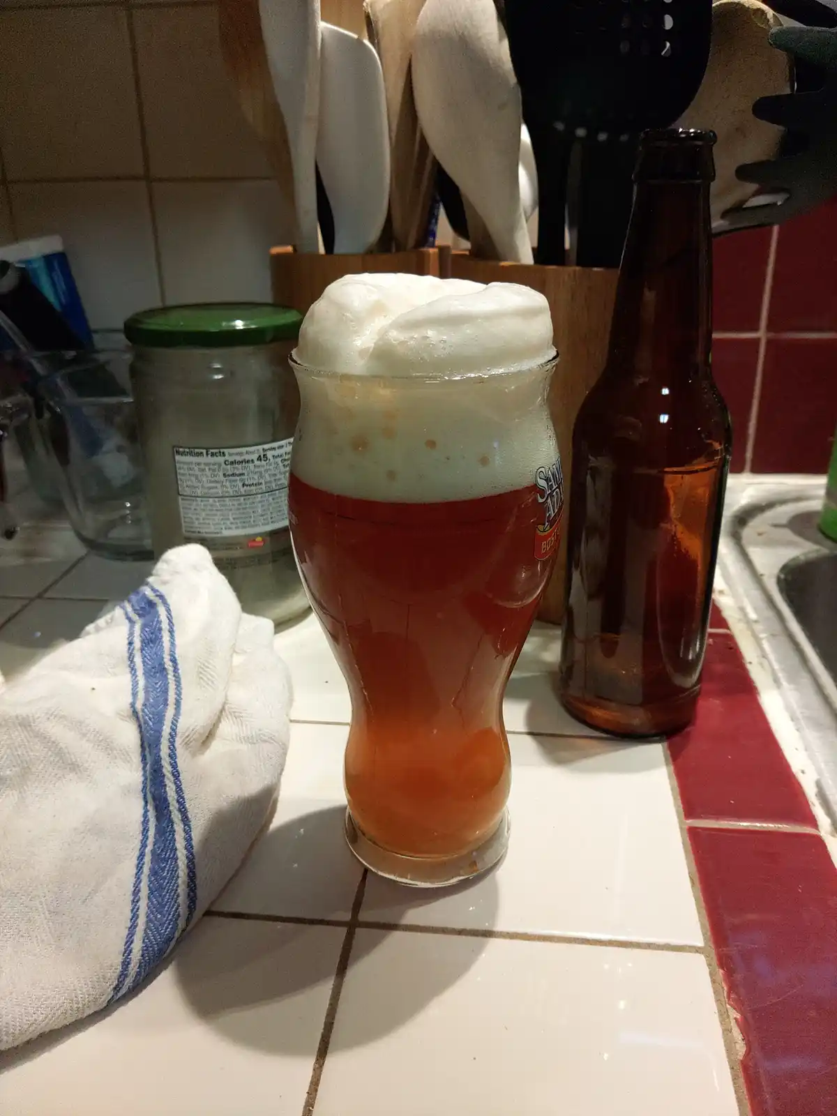 A glass of amber beer with an excessive, overflowing white foam head sits on a kitchen counter next to an empty brown…