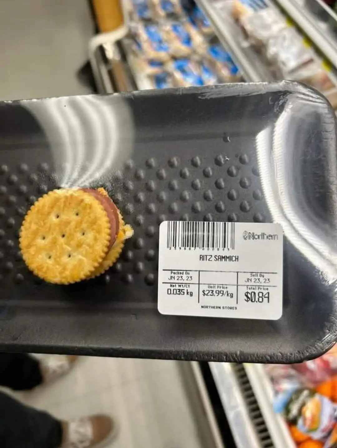 A single Ritz cracker sandwich with a slice of deli meat sits on a black plastic tray in a grocery store. The price tag…