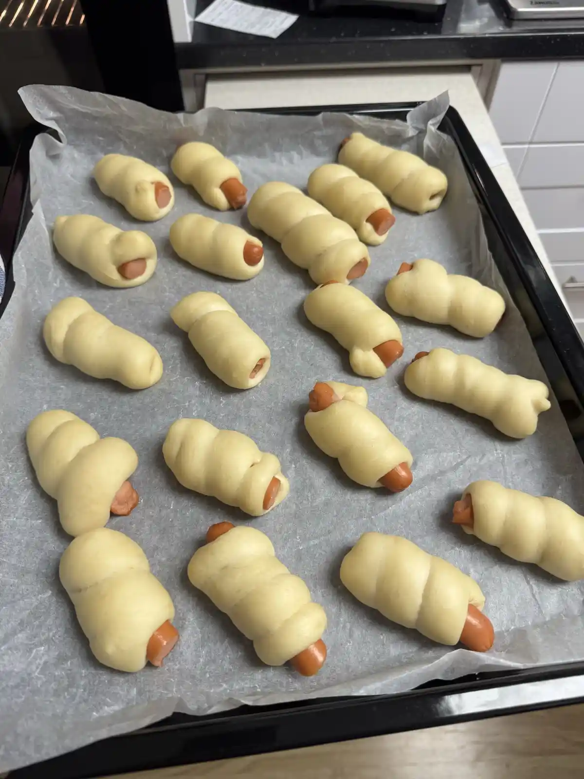 An overhead shot shows a baking tray lined with parchment paper, holding numerous uncooked pretzel rolls wrapped around hot…