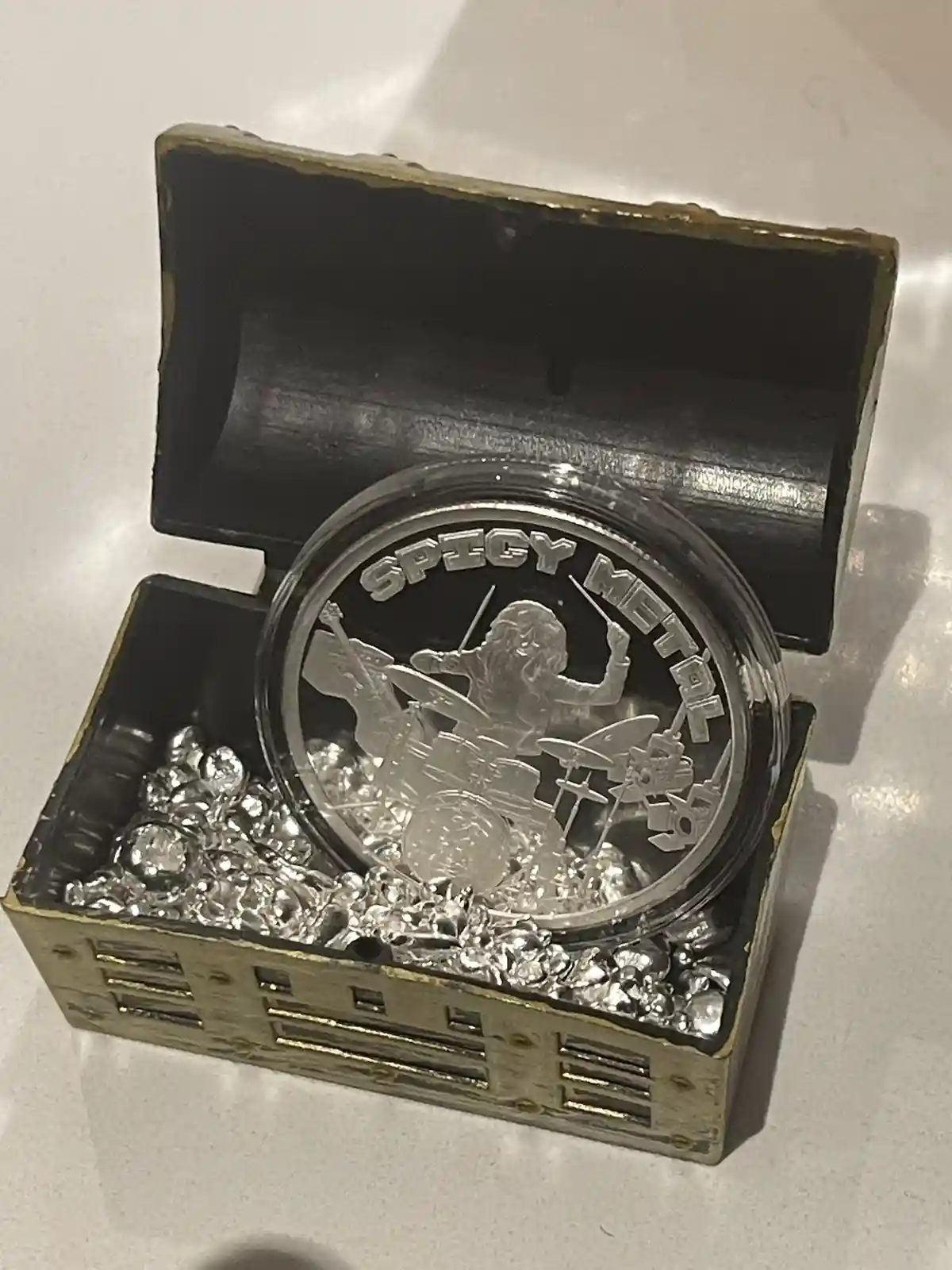A small, ornate treasure chest with a black interior and gold trim is open, revealing a large, clear circular medallion…