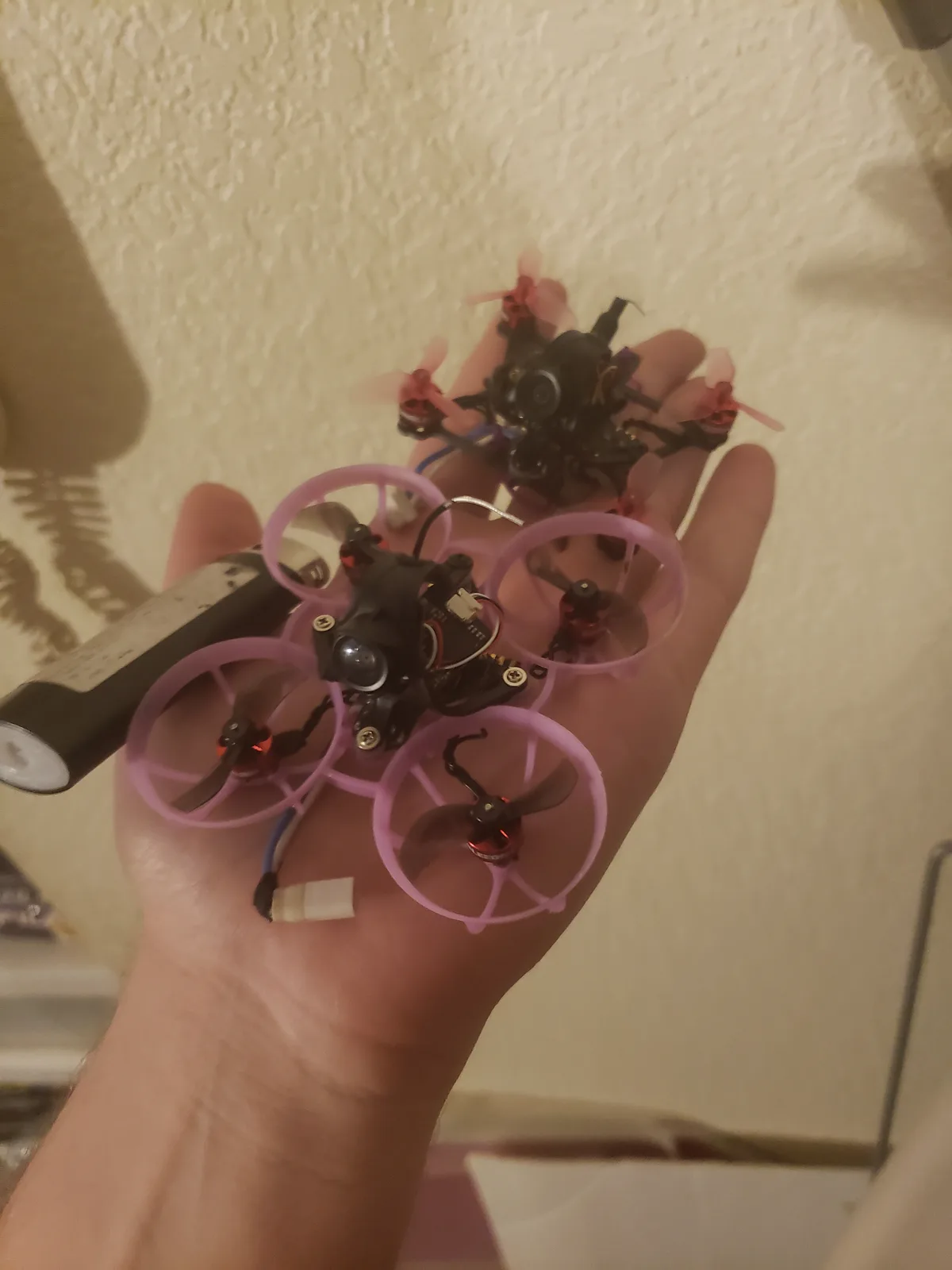 A person's hand holds two tiny quadcopters, one with pink protective frames and the other with red propellers, both with…