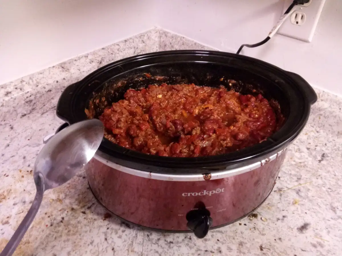 A reddish-brown chili sits in a black Crock-Pot on a speckled countertop, with a metal ladle resting on the side. The text…