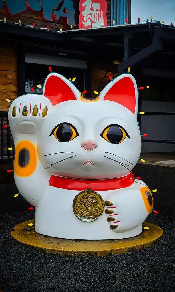 A large, white ceramic statue of a Maneki-neko (Japanese lucky cat) with red inner ears, yellow eyes, and a raised paw…