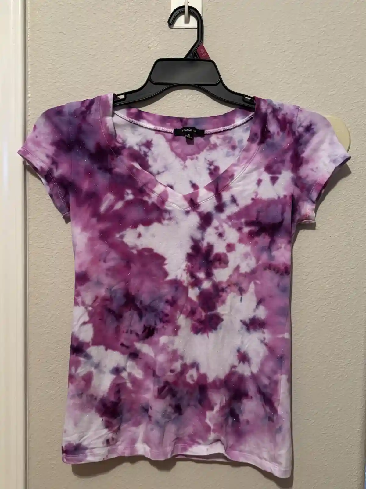 A purple and white tie-dye t-shirt with glitter, hanging on a black plastic hanger against a beige wall. The shirt has a…