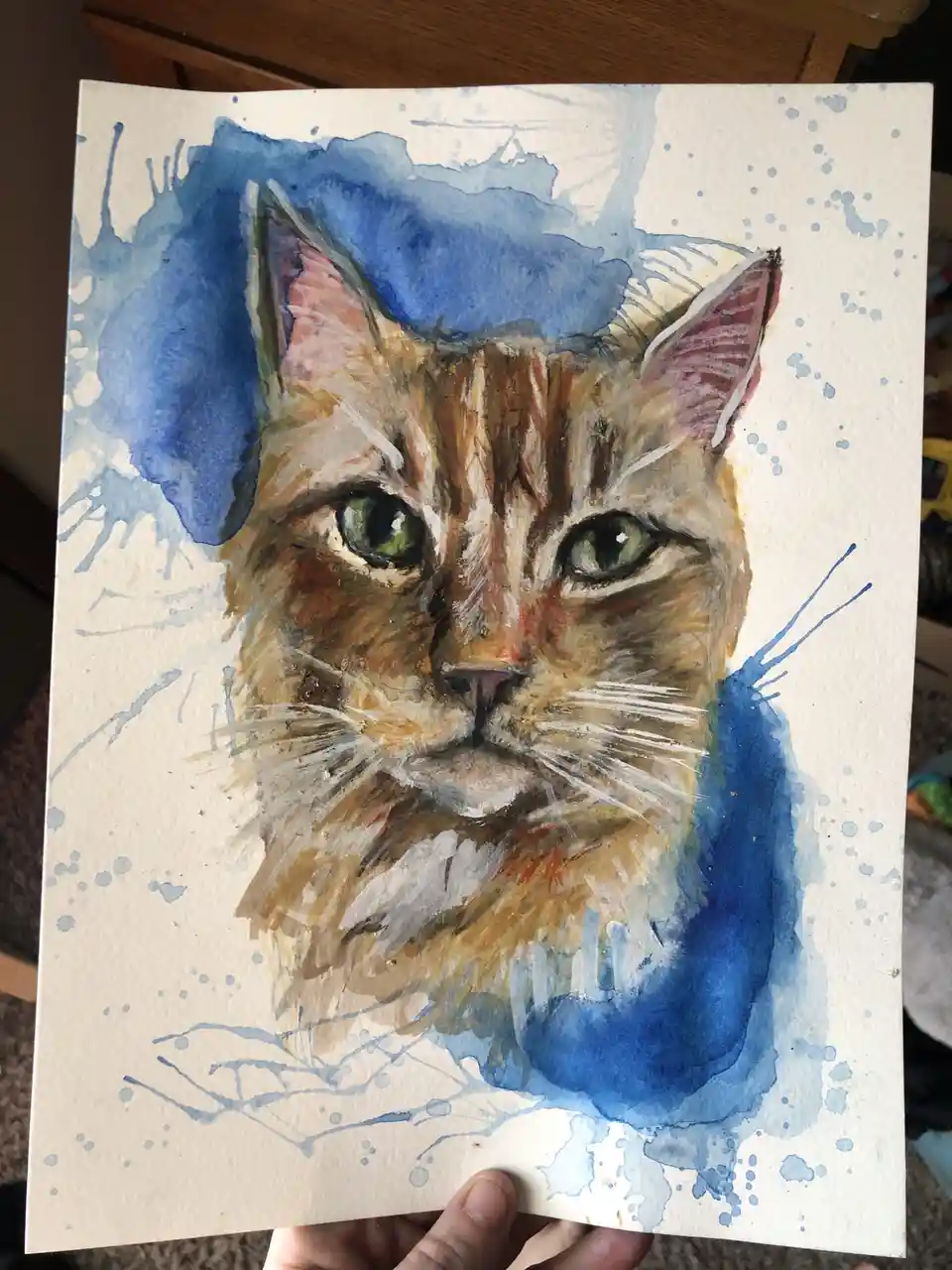 A watercolor painting of a cat's face with green eyes and brown fur, surrounded by blue paint splatters and washes. A hand…