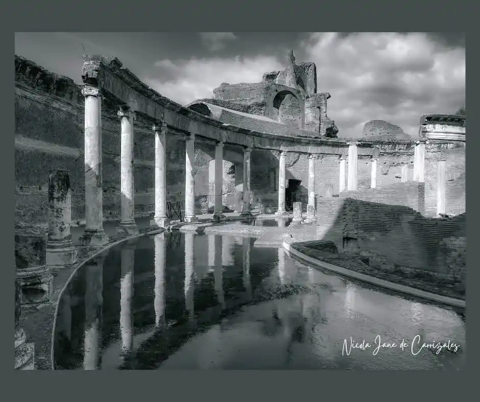 A black-and-white photograph of ancient Roman ruins with columns and a reflective pool of water under a cloudy sky. The…