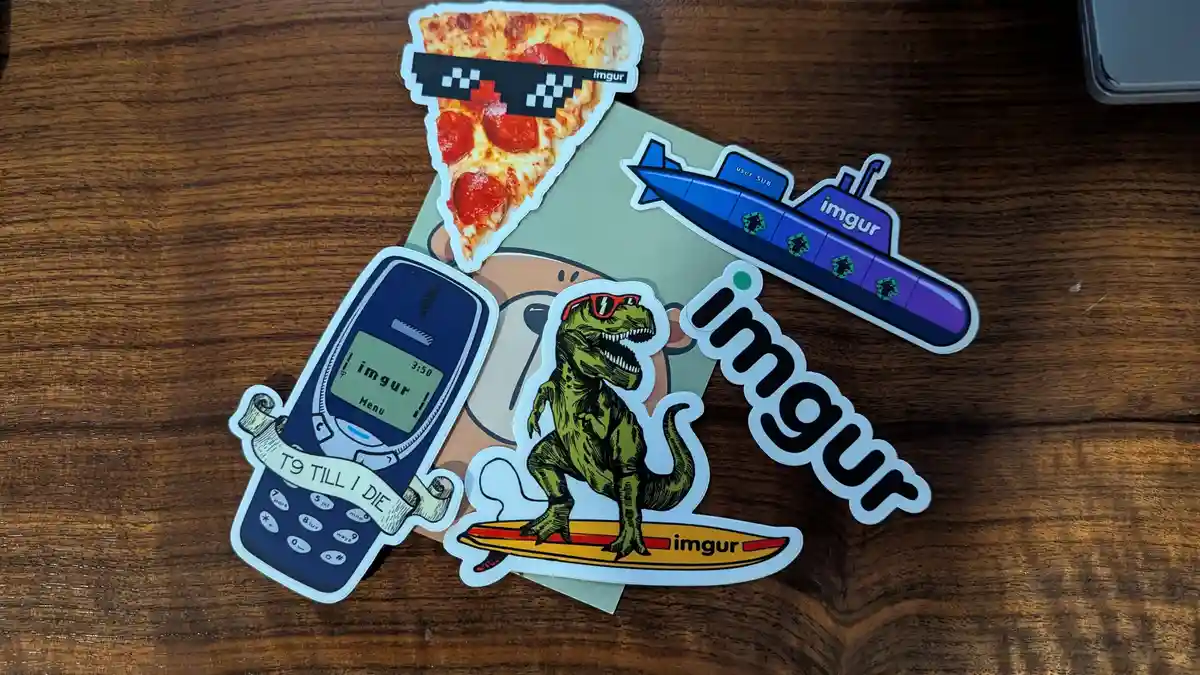 Several colorful Imgur-branded stickers are arranged on a wooden surface, featuring a slice of pepperoni pizza wearing…