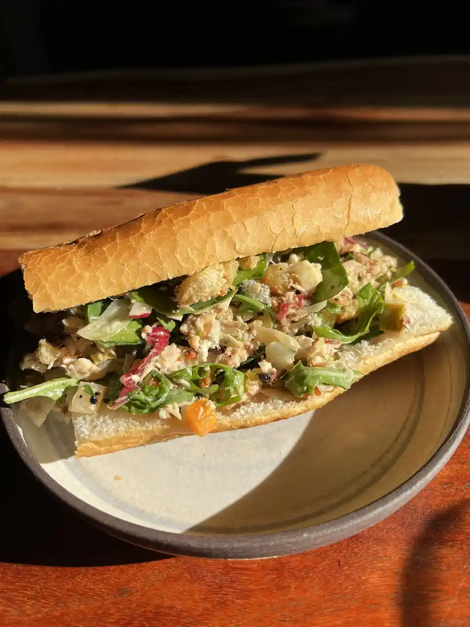 A close-up photo of a sandwich in a crusty baguette, filled with shredded chicken, greens, croutons, and other salad-like…