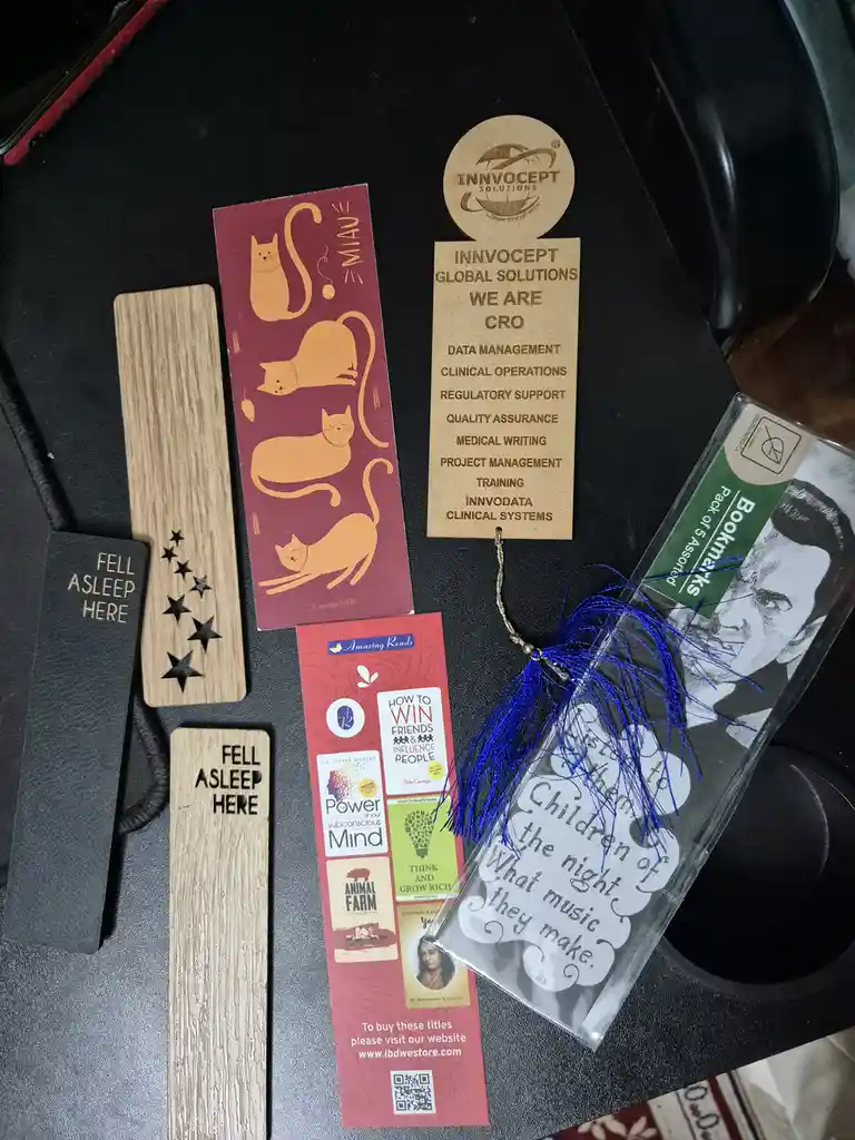 A collection of bookmarks laid out on a dark surface, including two wooden ones with 'FELL ASLEEP HERE' engraved, a red one…