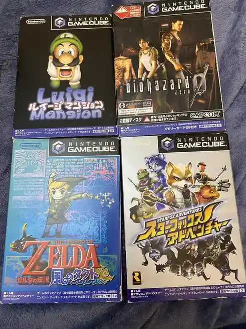 Four Japanese Nintendo GameCube game cases are laid out on a blue fabric surface. Top left is Luigi Mansion featuring Luigi…