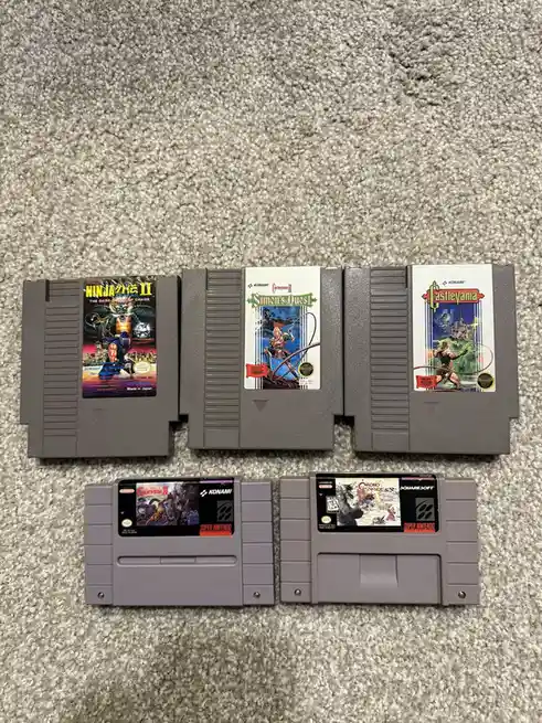 Five Super Nintendo cartridges are laid out on a gray carpet. Three are older, gray NES cartridges with labels for Ninja…