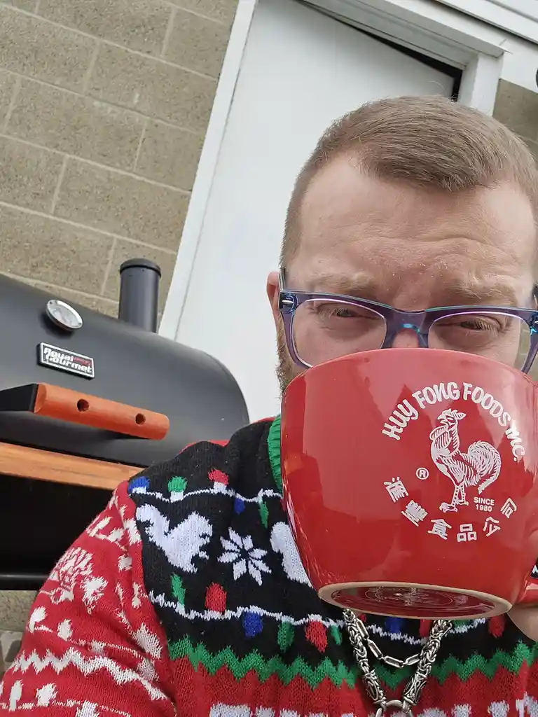 A man wearing a festive, colorful Christmas sweater and glasses holds a large red mug with the Huy Fong Foods logo…