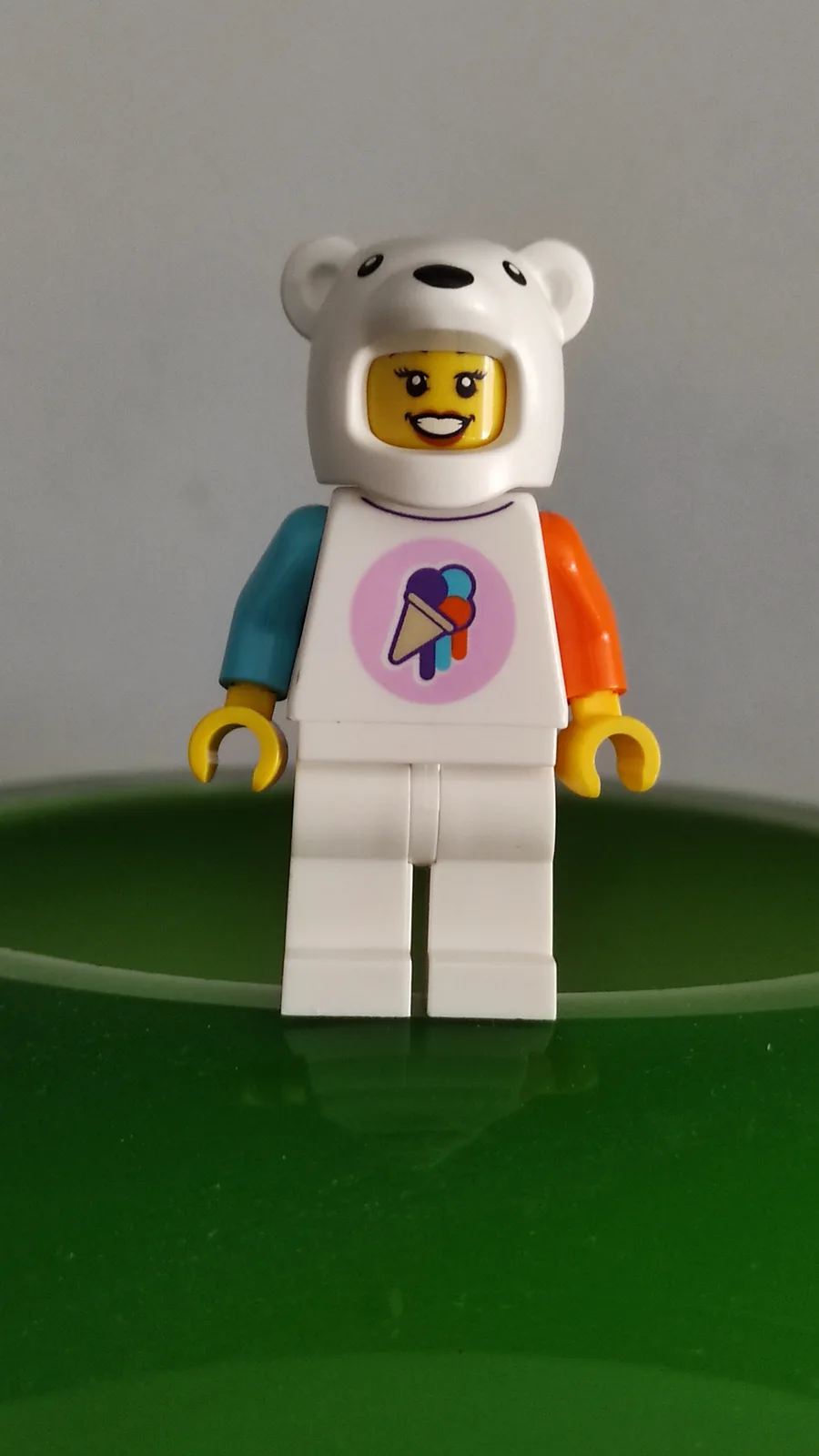 A lego minifigure with a yellow face and smiling expression wears a white polar bear hood and a white apron with a colorful…
