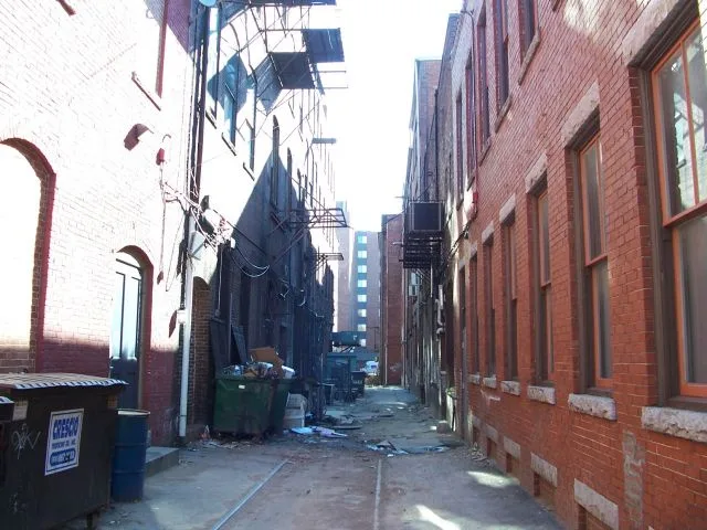 A narrow alleyway between red brick buildings with dumpsters, scaffolding, and debris. Overlaid text reads 'i'm not lazy i'm…
