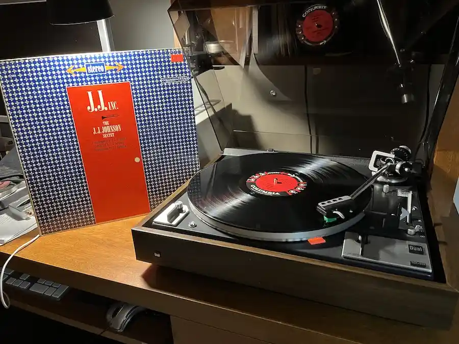 A vintage Dual turntable with a vinyl record playing, next to the album cover for 'J.J. Inc. The J.J. Johnson Sextet' by…