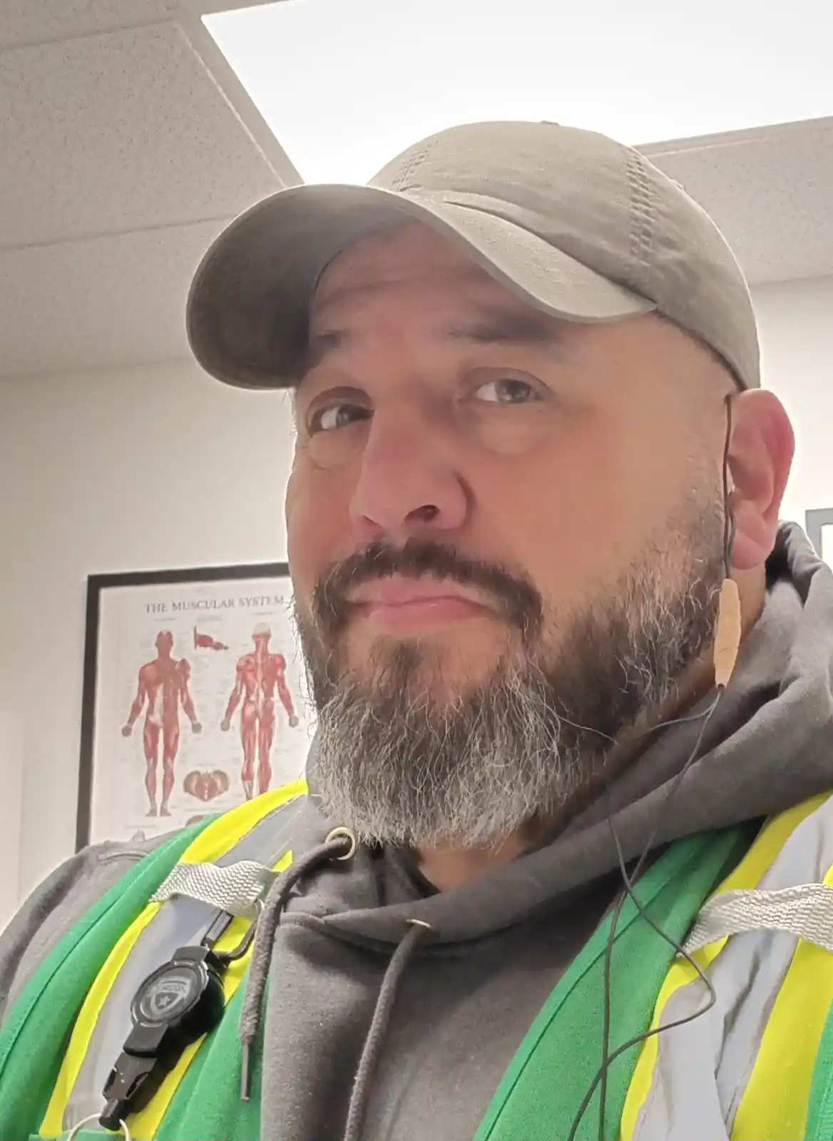 A man with a beard and cap, wearing a green and yellow safety vest over a hoodie, looks at the camera with a skeptical…