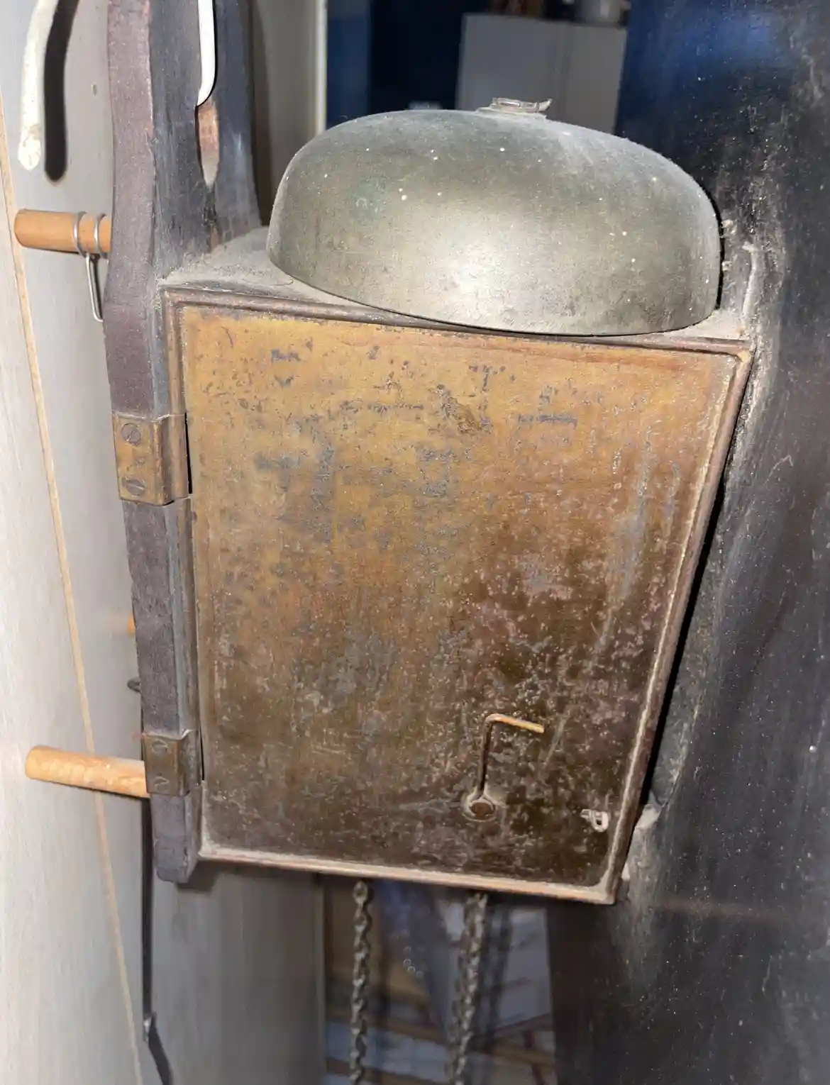 A close-up of a rusty, weathered, rectangular metal box with a dome-shaped lid, mounted on a dark surface. The box has a…