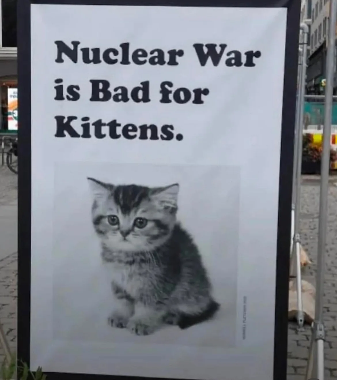 A large outdoor poster displays the text 'Nuclear War is Bad for Kittens.' above a black-and-white photograph of a small…
