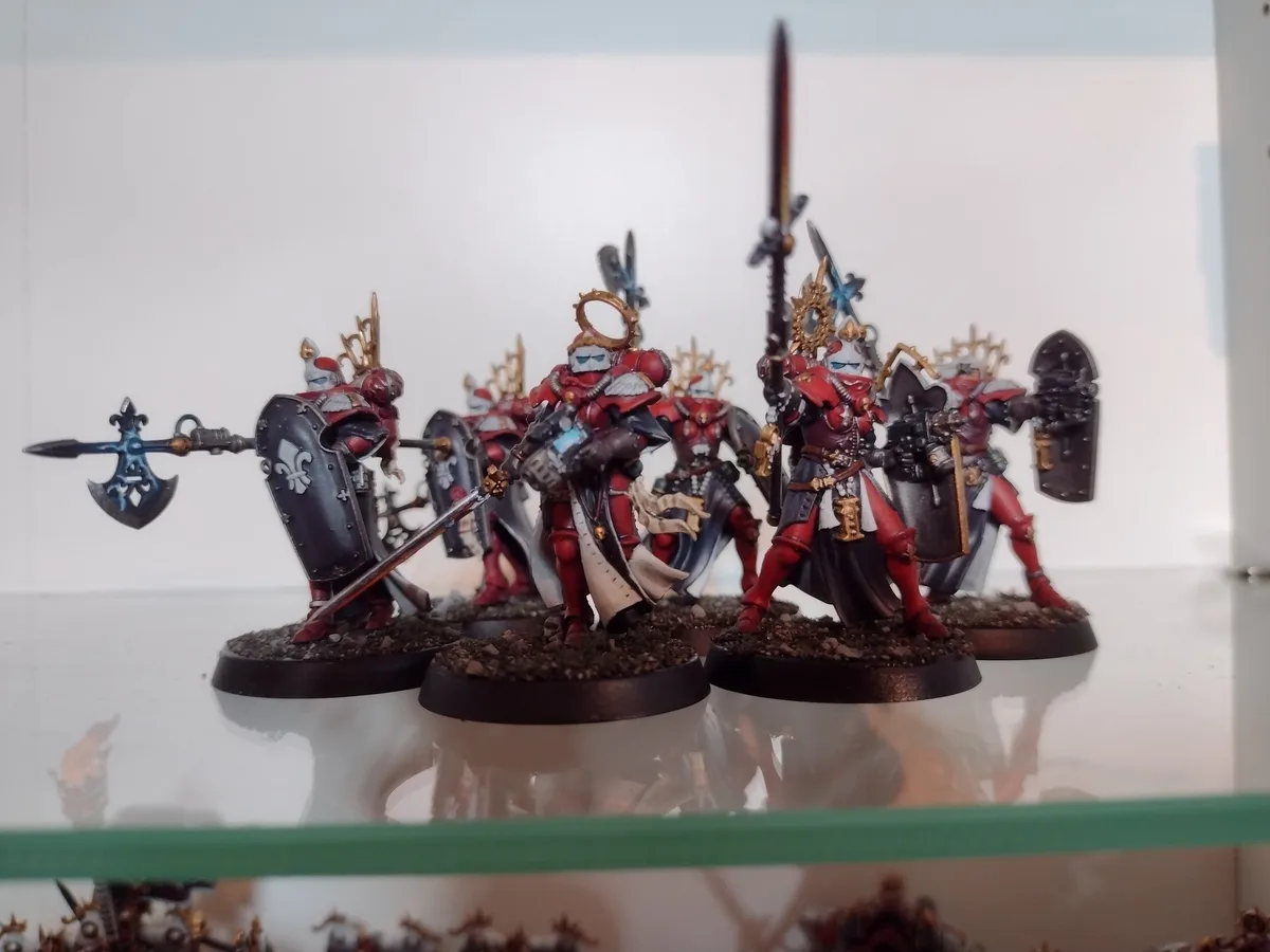 A row of intricately painted red and black fantasy miniatures, resembling Space Marines, are displayed on a glass shelf…