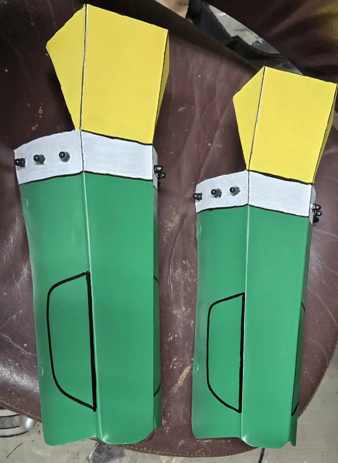 Two handmade, green and yellow arm guards with white bands and black bolts sit side by side on a brown leather surface. The…