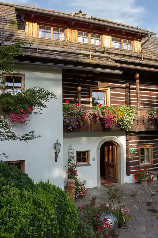 a picturesque, two-story wooden house with white stucco walls, a stone-paved courtyard, and vibrant flower boxes overflowing…