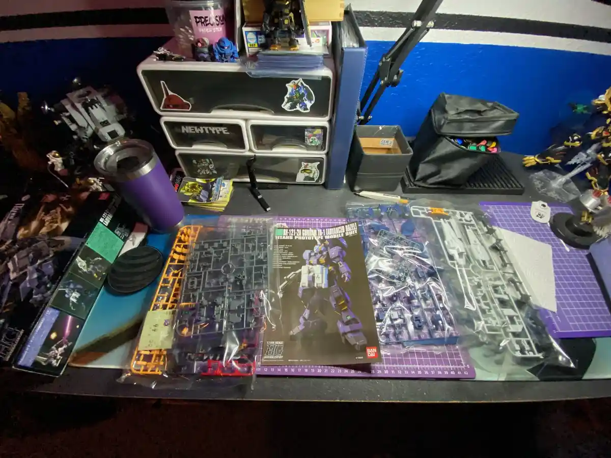 A cluttered hobbyist's desk is filled with Gundam model kits, parts, tools, and storage drawers, with the box for the…
