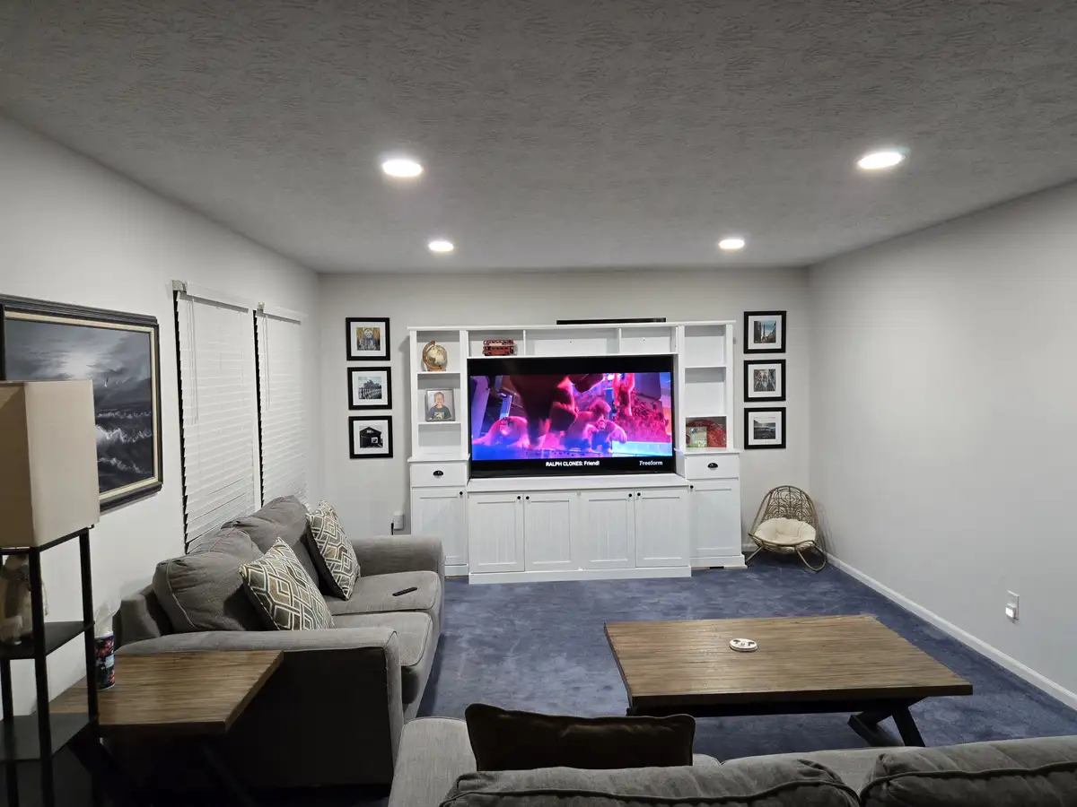 A well-lit, modern living room with gray sofas, a wooden coffee table, and blue carpeting. A large TV sits on a white media…